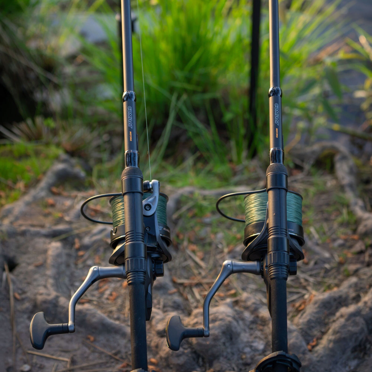 Two black Prologic COMBAT MICRO fishing rods with green line spools and silver handles resting on a rocky ground outdoors with blurred green grass in the background.
