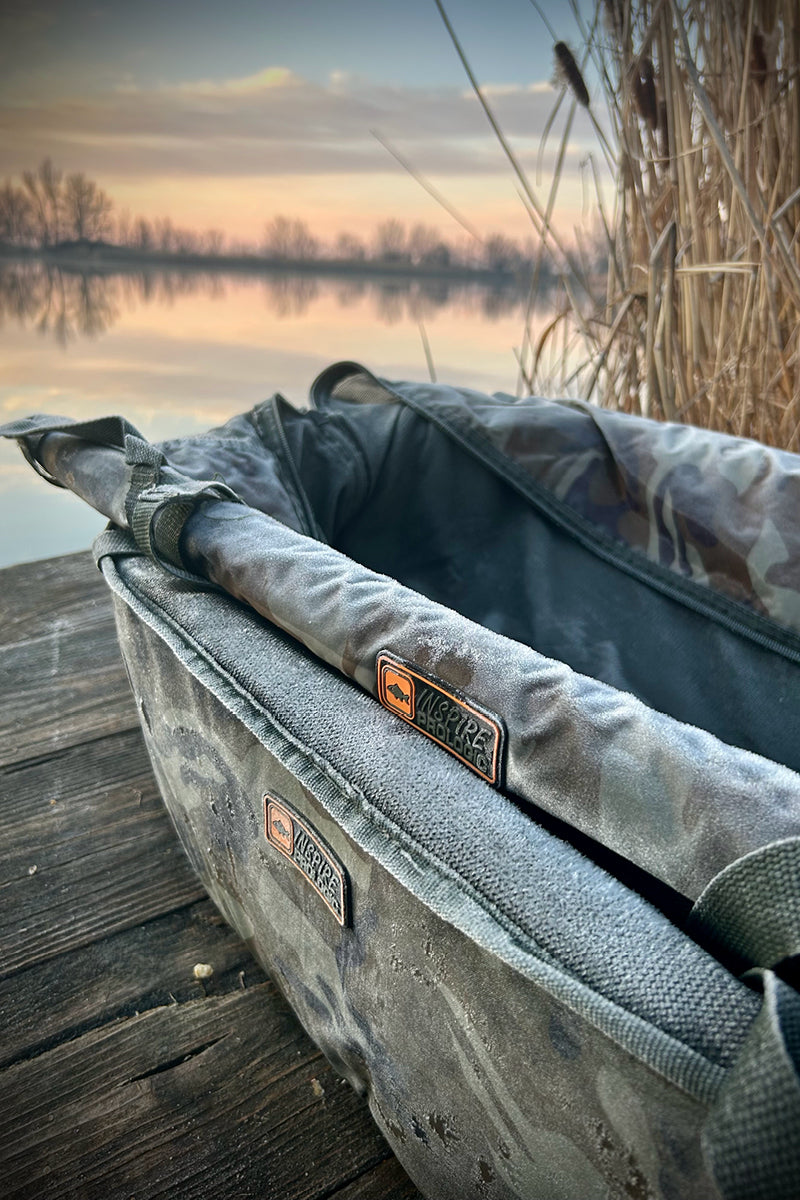 A camouflage Prologic Inspire unhooking mat, with rod holder on top, sits on a wooden dock with a lake and reeds in the background at sunset. 