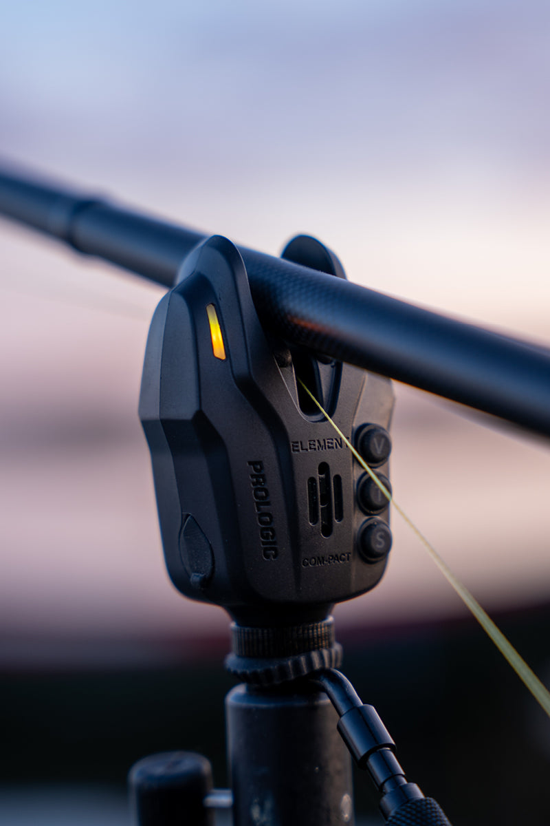 Close-up of a black Prologic Element fishing bite alarm with mounted rod and line, with a blurred outdoor background at dusk.