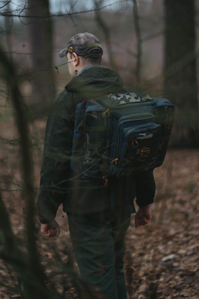 A man wearing a camouflage cap and dark jacket walks through a wooded forest while carrying a large dark blue and green outdoor backpack with orange zipper accents.