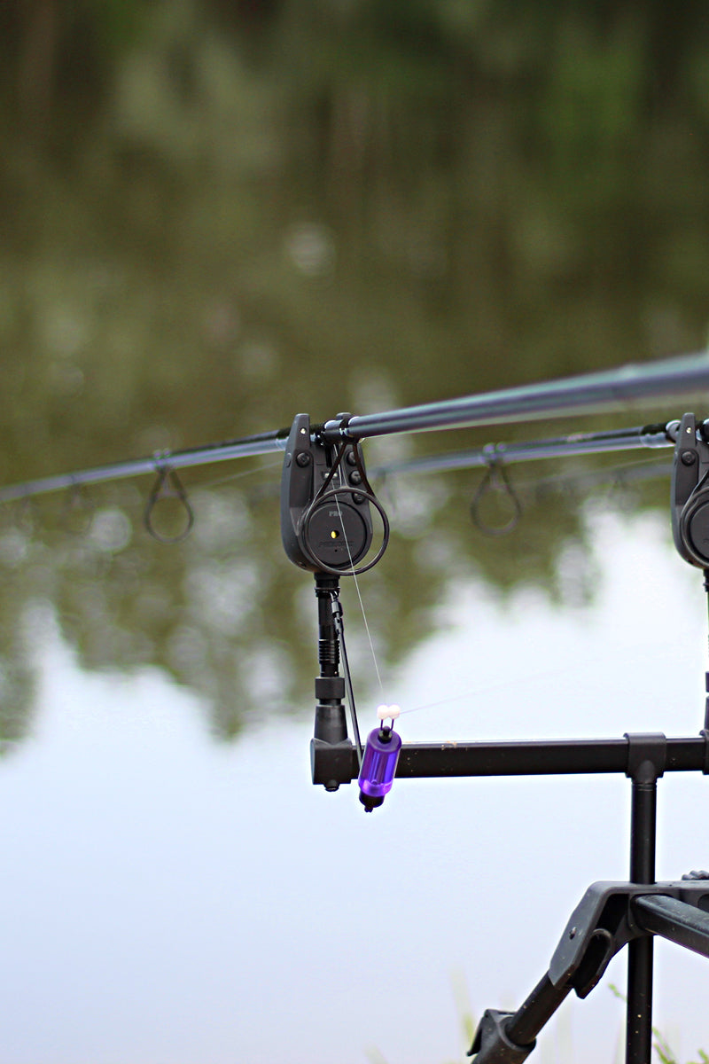 Close-up of a fishing rod mounted on a rod holder and bite alarm and indicator near a lake, with a purple weight attached to the line and a blurred natural background of water and trees.