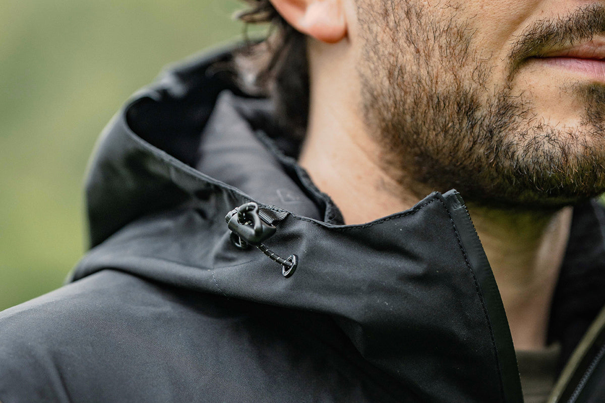 Close-up of a man wearing a black hooded jacket, showing the drawstring hood and chin strap detail against a blurred green outdoor background.