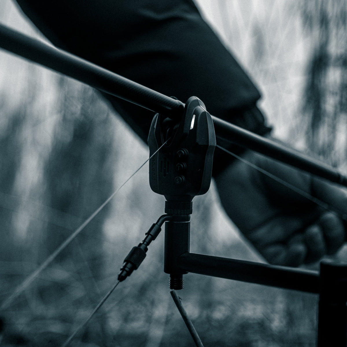 Close-up of a bite alarm sitting on a rod bar holding a rod. A person's arm and hand visible in a blurred outdoor setting