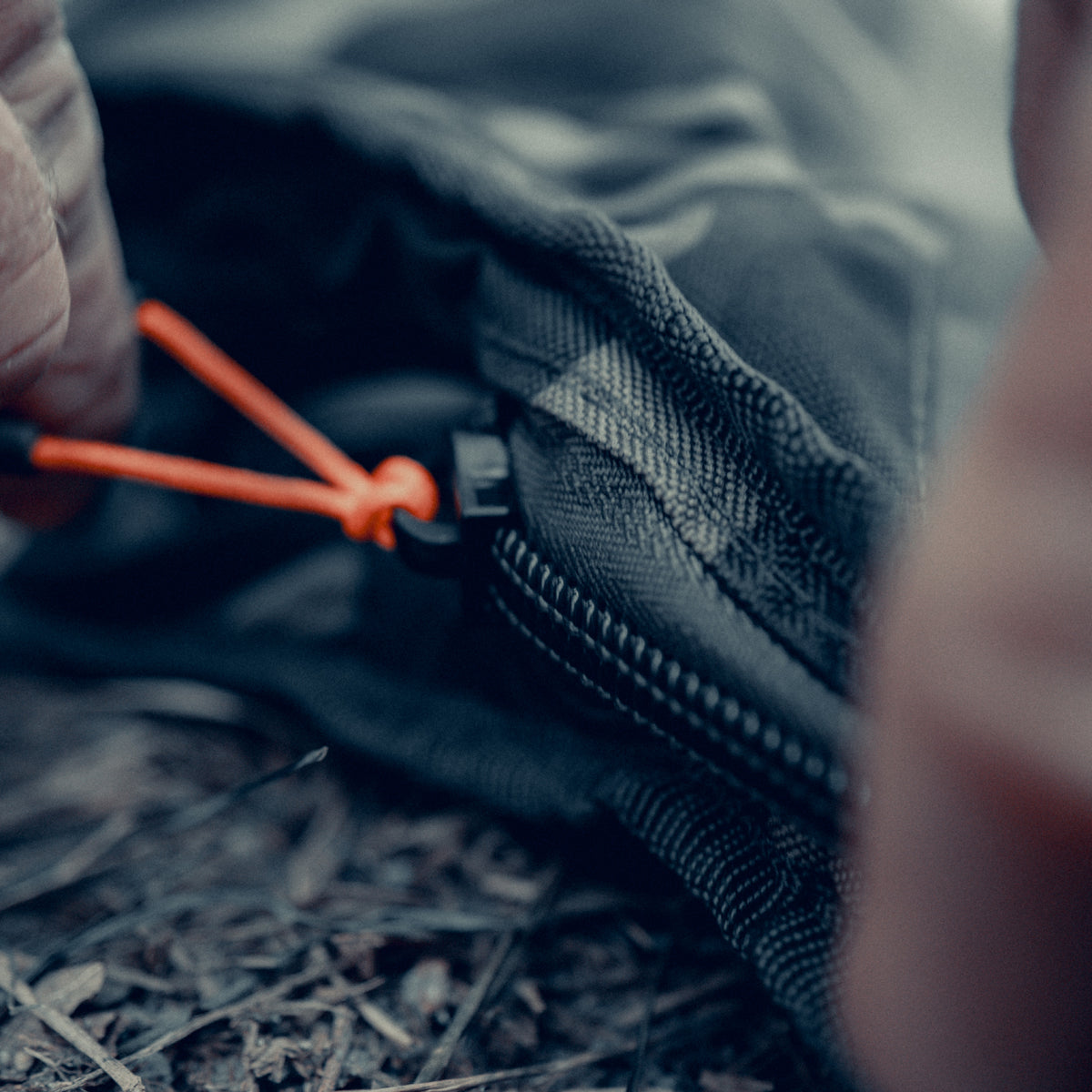 Close-up of a person's hand adjusting an orange drawstring zip puller on a black fabric bag, set against a blurred outdoor ground background.