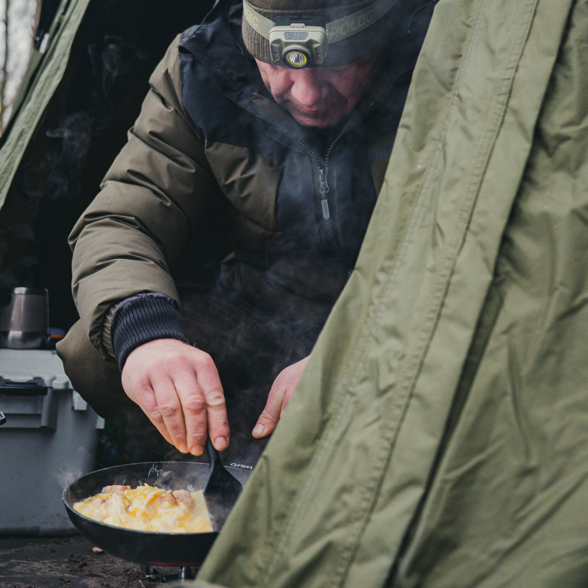 A man wearing a headlamp and a dark puffer jacket cooks food in a frying pan inside a green tent, with steam rising from the dish.